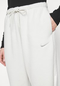 Valkoiset fleece-housut, joissa on elastinen vyötärö ja naru, sekä sivutaskut. Hopeinen Nike-logo on brodeerattu lähelle lantiota.