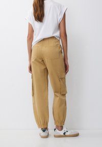Khaki spodnie cargo z elastycznymi mankietami, kieszeniami po bokach i luźnym krojem. Noszone z białą bluzką z krótkim rękawem i trampkami.