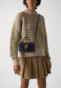 Stripet strikkegenser i gul og grå, kombinert med et beige plisserte skjørt. En liten svart håndveske med gullhardware henger fra skulderen.