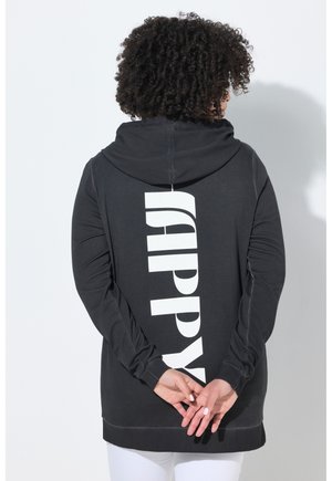 Personne aux cheveux bouclés portant un sweat à capuche noir avec le texte vertical blanc "HAPPY" dans le dos, mains jointes derrière, pantalon blanc.