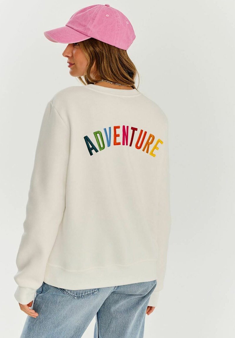 Bílá mikina s barevným výšivkou "ADVENTURE" na zadní straně, doplněná růžovou baseballovou kšiltovkou. Má uvolněný střih a žebrované manžety.
