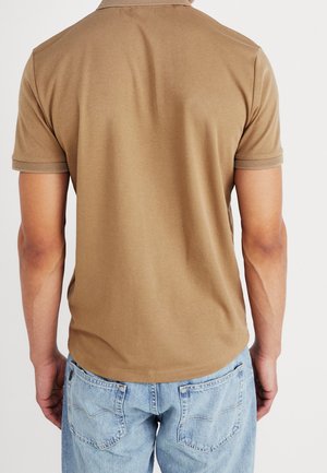 Mann trägt ein schlichtes braunes Polohemd mit kurzen Ärmeln und hellblaue Jeans, von hinten vor einem einfarbigen Hintergrund gezeigt.