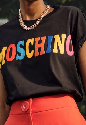 Person trägt ein schwarzes T-Shirt mit buntem "MOSCHINO"-Schriftzug und eine klobige goldene Kettenhalskette, kombiniert mit hoch taillierten roten Hosen mit einem großen Knopf.