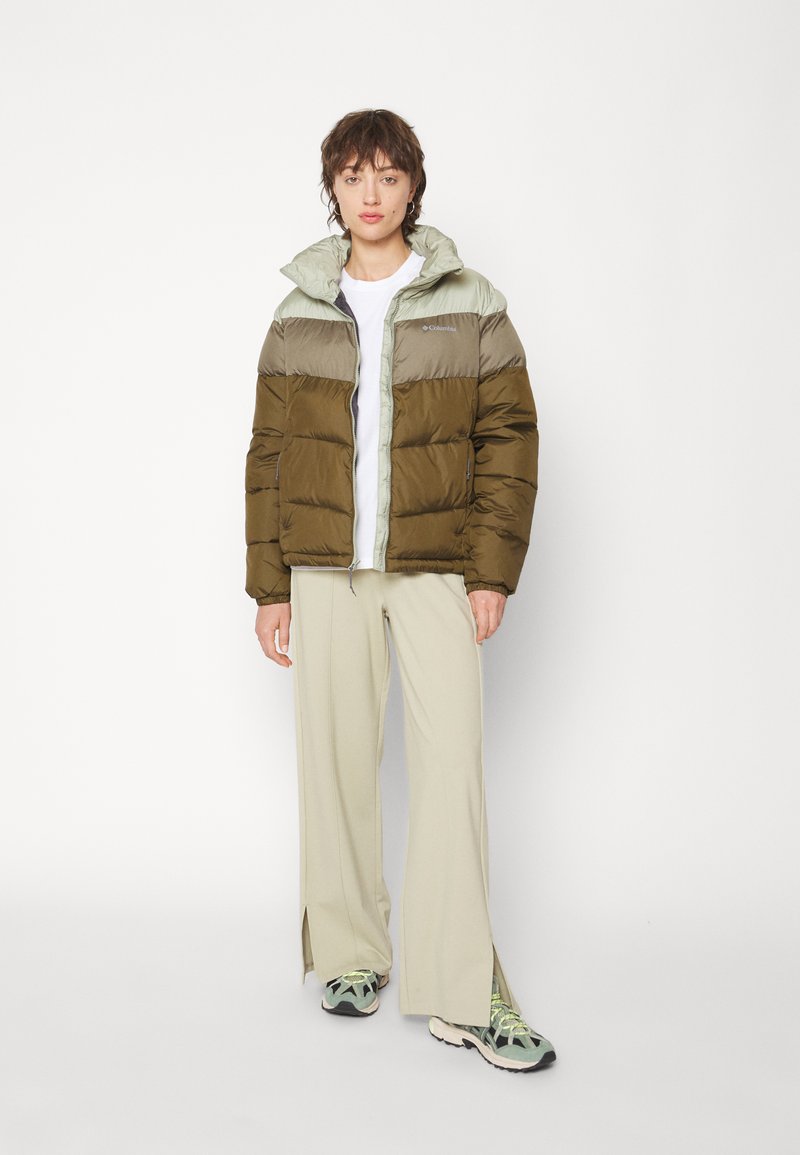 Chaqueta tipo pluma en verde oliva y gris claro, con un cuello alto y cierre de cremallera. El modelo lleva pantalones anchos beige y zapatillas.