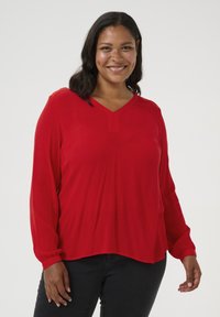 Blusa roja de manga larga con escote en V y sutil textura de rayas verticales. La tela es suave y el dobladillo está ligeramente redondeado en la parte inferior.
