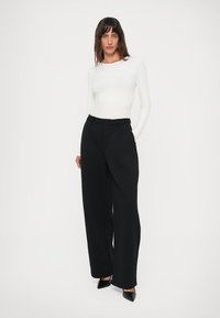 Woman wearing a white long-sleeve top, high-waisted black wide-leg trousers, and black pointed-toe heels, standing against a white background.