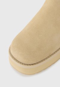 Nærbilde av beige semsket skotupp med synlig søm og tykk gummisåle på en hvit bakgrunn.