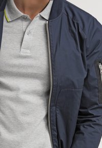 Marinblå bomberjacka med dragkedja framtill, ribbad krage och ficka på vänster ärm. Bärs över en ljusgrå polo med krage.