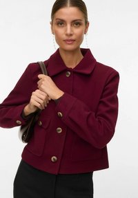 Frau mit nach hinten gekämmtem Haar, die lange Kettenohrringe trägt, eine burgunderrote Jacke mit Knöpfen und eine braune Handtasche über der Schulter hält.