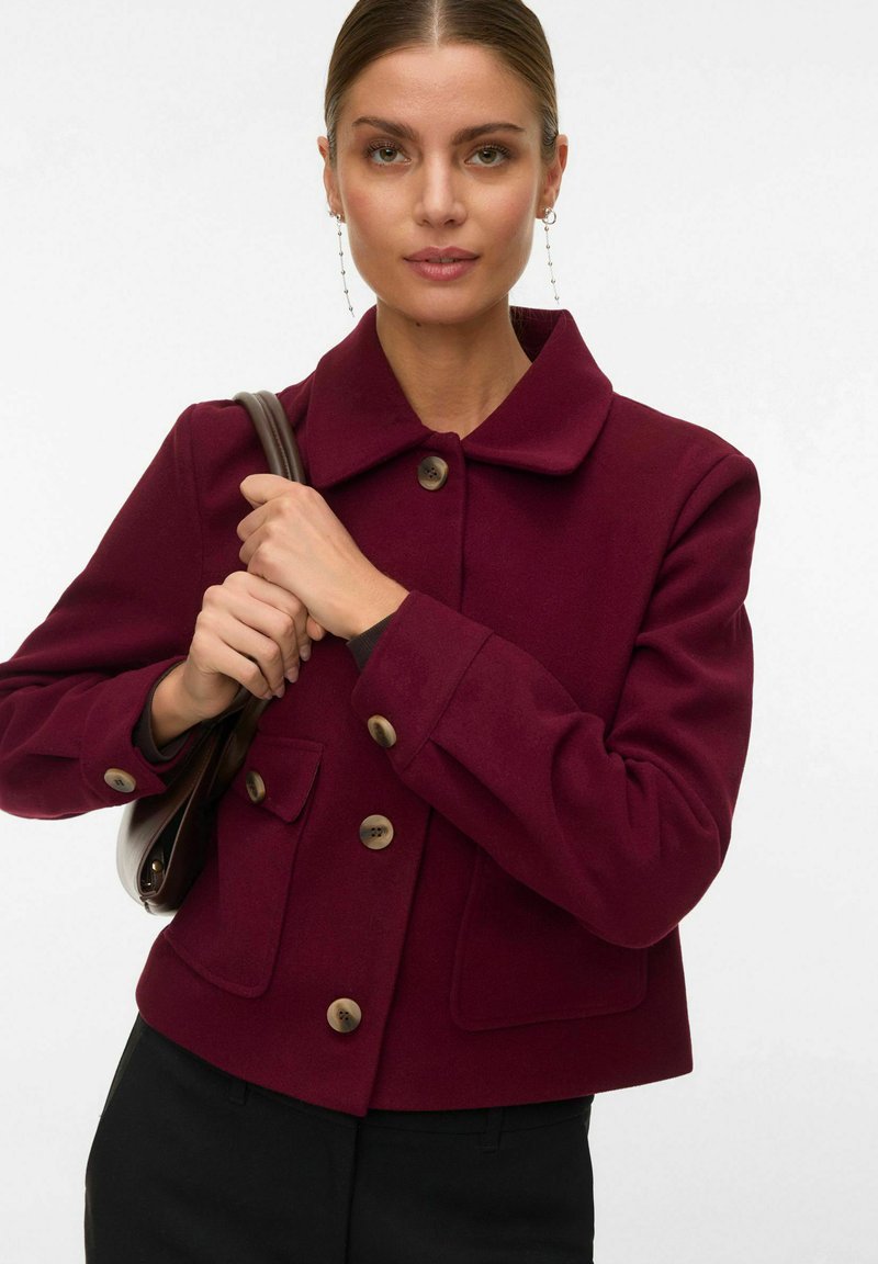 Frau mit nach hinten gekämmtem Haar, die lange Kettenohrringe trägt, eine burgunderrote Jacke mit Knöpfen und eine braune Handtasche über der Schulter hält.