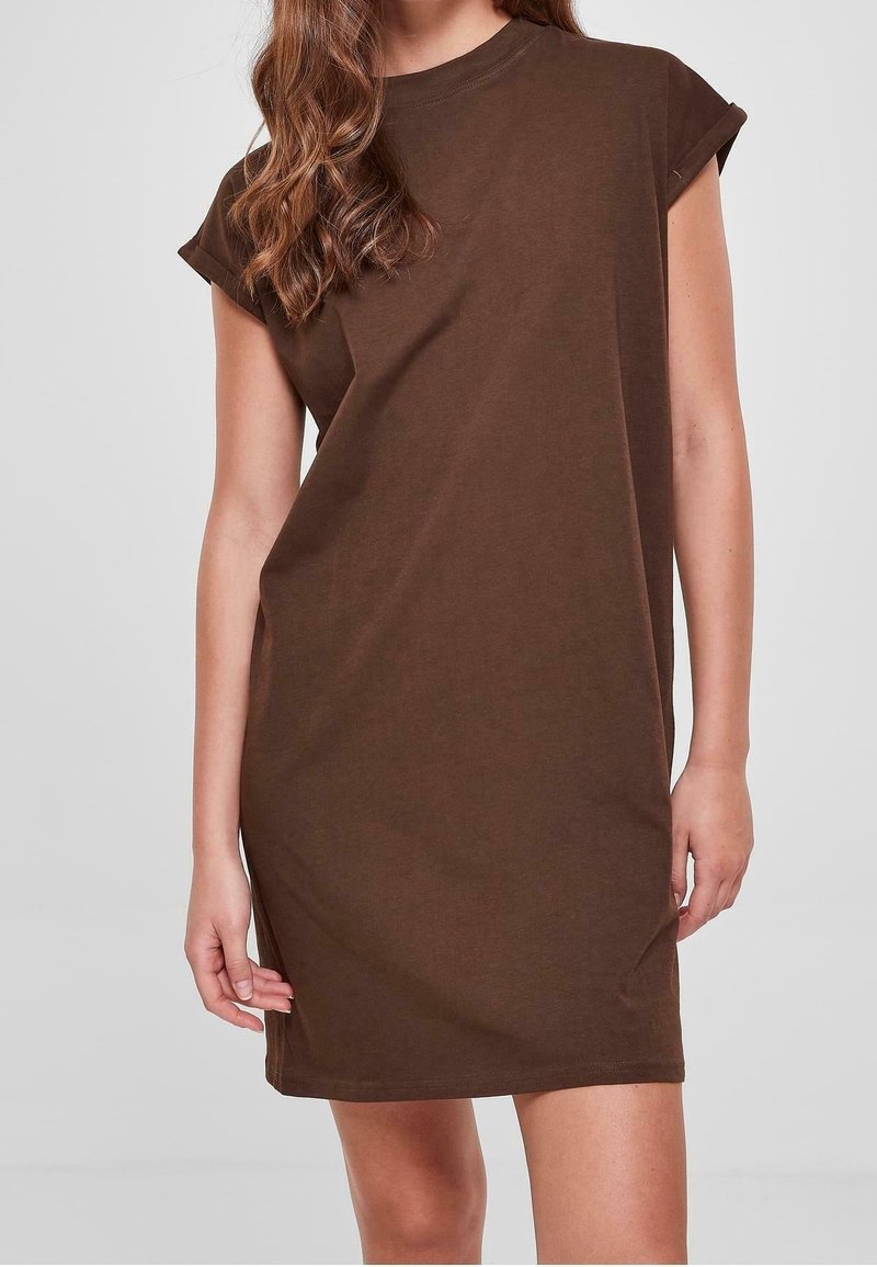 Femme portant une robe T-shirt marron à manches courtes, ample, debout devant un fond gris clair uni.