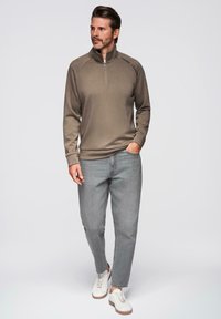 Sudadera con cremallera de color marrón claro y cuello alto, con texto negro y puños de canalé, combinada con jeans claros ajustados y zapatillas blancas.