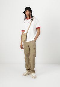 White t-shirt with red collar, beige cargo pants with side pockets, black bucket hat, and beige crossbody bag. Neutral background.