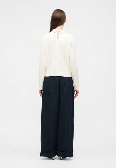 Femme aux longs cheveux bruns, portant un pull blanc côtelé avec une ouverture en forme de goutte dans le dos et un pantalon bleu marine évasé avec des poches plaquées, debout de dos.