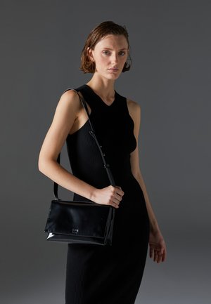 Black patent leather handbag with a structured shape, featuring a detachable shoulder strap and a glossy finish. Styled with a ribbed black dress.