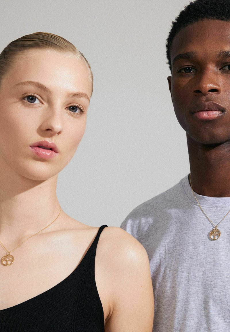 Des colliers en or avec des pendentifs circulaires portés par un homme et une femme. La femme porte un débardeur noir ; l'homme a un t-shirt gris. Textures lisses.