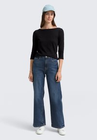 Schwarzes langärmliges Oberteil, hoch taillierte weite blaue Jeans, weiße Sneaker und eine hellblaue Mütze, vor einem neutralen Hintergrund präsentiert.