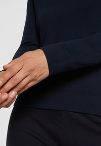 Mains ajustant le poignet d'un pull bleu marine à manches longues sur un pantalon bleu marine assorti, sur un fond gris clair uni.