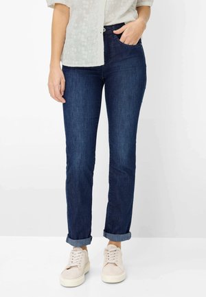 Donna con jeans blu scuro risvoltati, camicia bianca a fiori e sneakers bianche, in piedi con una mano in tasca su sfondo bianco.