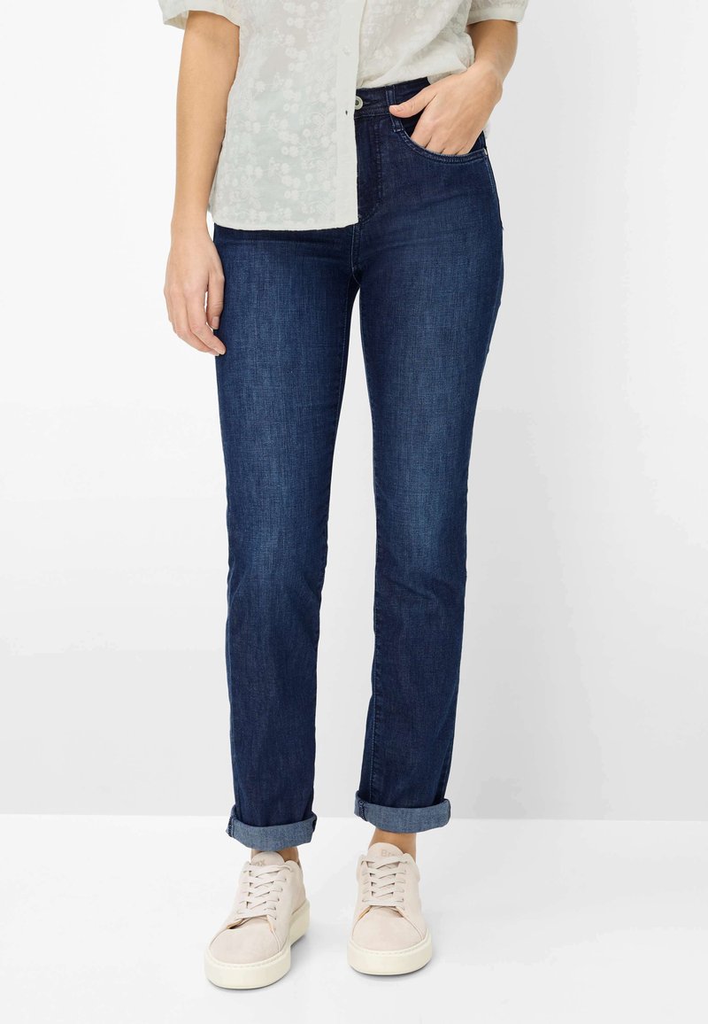 Frau trägt umgeschlagene dunkelblaue Jeans, weißes Blumenhemd und weiße Sneaker, steht mit einer Hand in der Tasche vor weißem Hintergrund.