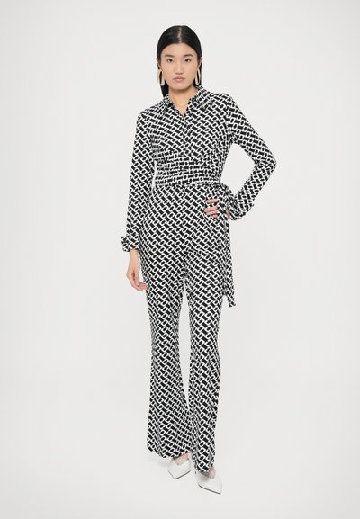 Μαύρη και λευκή τυπωμένη ολόσωμη φόρμα με μακριά μανίκια, σχέδιο τύπου wrap, παντελόνι με φαρδιά πόδια και λεπτομέρεια δέσιμου στο πλάι. Λείο ύφασμα με γεωμετρικό μοτίβο.