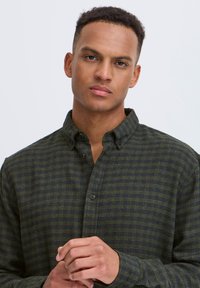 Ung mand med kort krøllet hår iført en grøn og marineblå ternet skjorte, stående mod en simpel lys baggrund.