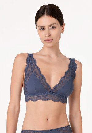 Donna che indossa un bralette di pizzo blu con motivi floreali e bordi smerlati, in piedi davanti a uno sfondo bianco uniforme.
