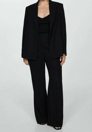 Kobieta ubrana w czarną, dopasowaną marynarkę, czarny top z dekoltem w serek oraz szerokie czarne spodnie stojąca na tle jasnej, jednolitej ściany.