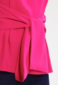 Blusa fucsia con design a portafoglio e fiocco annodato sul davanti, realizzata in tessuto morbido, con una vestibilità ampia e dettagli laterali arricciati.