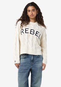 Baltas megztinis su pynimo raštu, su „REBEL“ tekstu tamsiai mėlynos spalvos. Megzti манжети ir apačia, derinamas su mėlynomis džinsomis.