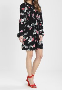 Svart blommig klänning med långa ärmar, med rosa och gröna blommönster, knäppt midja och utåtvänd fåll; i kombination med röda klackar.