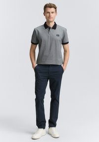 Grå polo-shirt med sort krave og manchetter, parret med mørkeblå bukser og hvide sneakers. Modellen står med hænderne i lommerne.