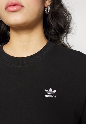 Femme portant un sweat-shirt Adidas noir avec un logo blanc, des boucles d'oreilles argentées en anneau et des cheveux ondulés foncés visibles au-dessus du col.