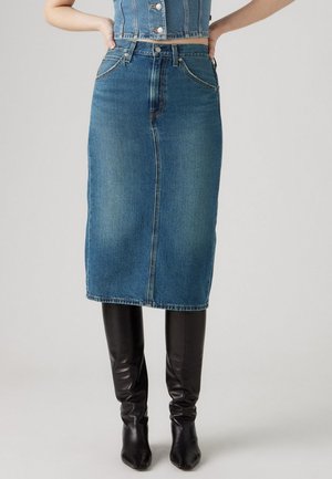 Denim blyantkjole i mellomblått med høy midje, rett snitt, sentral søm foran og fem lommer. Kombinert med høye svarte støvler.