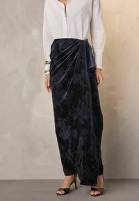 Dunkelblauer Samtrock mit floralem Stickerei, mit einem drapierten Design, kombiniert mit einem weißen Hemd und schwarzen offenen Pumps.