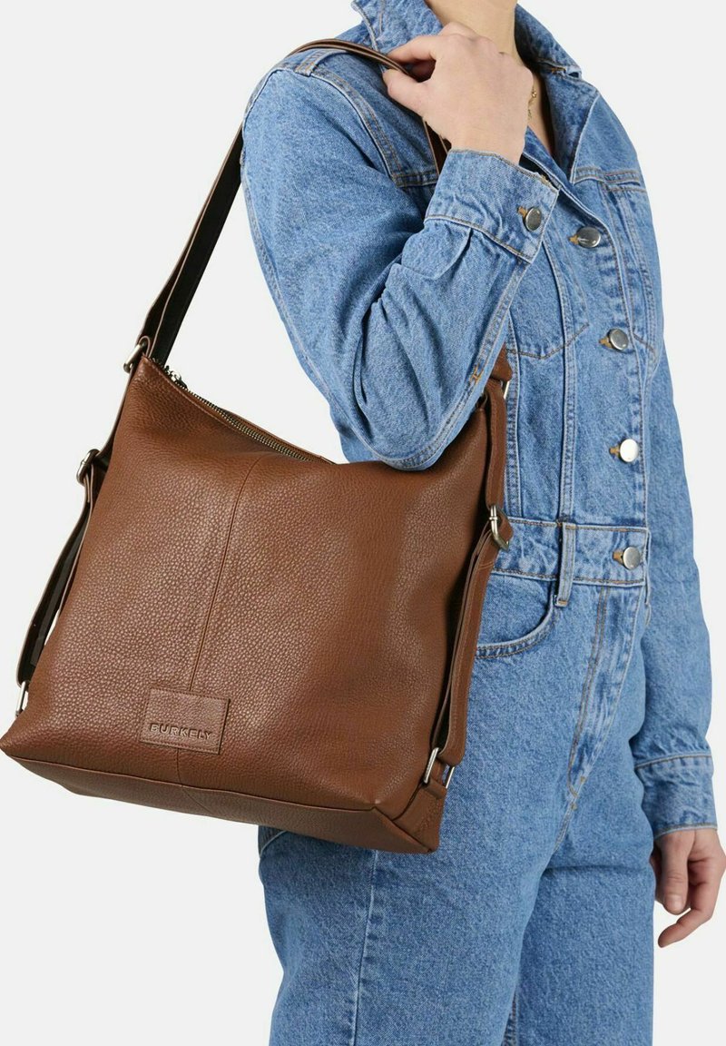 Bolso de hombro de cuero marrón con superficie texturizada, forma rectangular, herrajes de plata y correa ajustable, llevado con un conjunto de denim.
