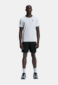 Vit sporttshirt med logotyp, i kombination med svarta shorts. Modellen bär grå löparskor och vita strumpor, stående mot en neutral bakgrund.