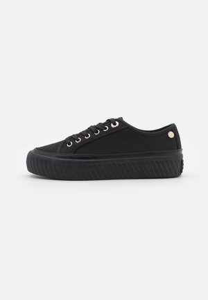 Czarny niski sneaker z grubą fakturowaną gumową podeszwą, materiałową cholewką, czarnymi sznurowadłami przez metalowe przelotki i małym, okrągłym logo blisko pięty.