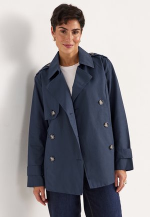 Woman with short dark hair wearing a navy double-breasted trench coat and dark jeans, standing against a plain light background.