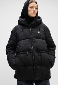 Mustapuffer takki vetoketjulla, säädettävällä hupulla ja teksturoidulla kankaalla. Oikeassa rinnassa on pieni logo.