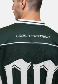 Donkergroen netstof voetbalshirt met witte accenten. Kenmerkt de tekst "GOODFOR NOTHING" geborduurd in het wit op de achterkant, met contrasterende ribbelkraag.