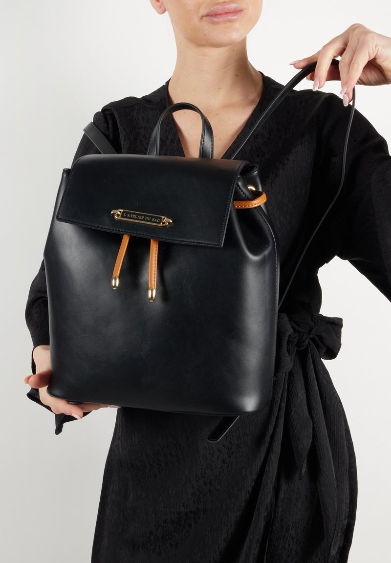 Musta nahkainen reppu, jossa on sileä pinta, etupaneeli, kullanväriset yksityiskohdat ja oranssit nahkaelementit. Varustettu säädettävillä hihnoilla ja minimalistisella muotoilulla.