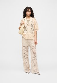 Γυναίκα που φοράει μπεζ πλεκτό τοπ και παντελόνι, κρατώντας μια μπεζ τσάντα, στέκεται μπροστά σε απλό λευκό φόντο.