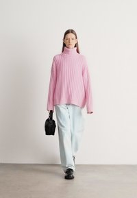 Přerostlý růžový rolák s žebrovaným vzorem ve spojení s světle modrými širokými kalhotami a černou obuví, držící malou černou kabelku.