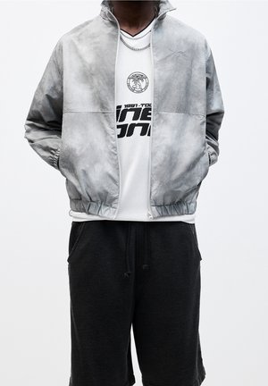 Personne portant une veste zippée grise par-dessus un t-shirt blanc à motif graphique et un pantalon de jogging noir, les mains dans les poches de la veste.