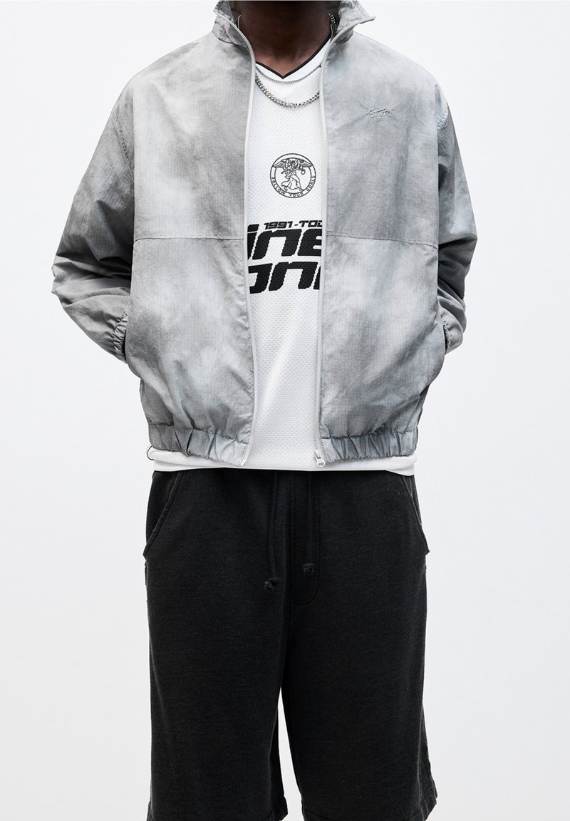 Personne portant une veste zippée grise par-dessus un t-shirt blanc à motif graphique et un pantalon de jogging noir, les mains dans les poches de la veste.