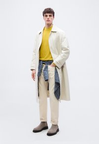 Jeune homme debout portant un trench-coat beige, un pull moutarde, un pantalon crème avec une chemise à carreaux bleus nouée à la taille, et des bottes grises sur fond blanc.
