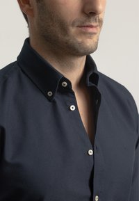 Camisa de botonadura en azul marino con una textura suave, que presenta un cuello clásico, botones blancos y un pequeño logo en el lado izquierdo.