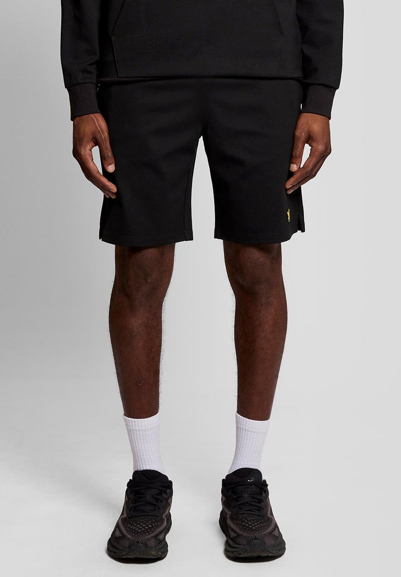 Homme portant un short noir, des baskets noires, des chaussettes blanches et un haut à manches longues noir, debout devant un fond gris uni.