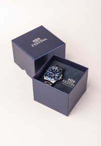 Mėlynas Festina laikrodis su sidabriniais akcentais, pateiktas tamsiai mėlyname dėkle su minkštu vidumi. Yra apvalus ciferblatas, nerūdijančio plieno apyrankė ir žymekliai.